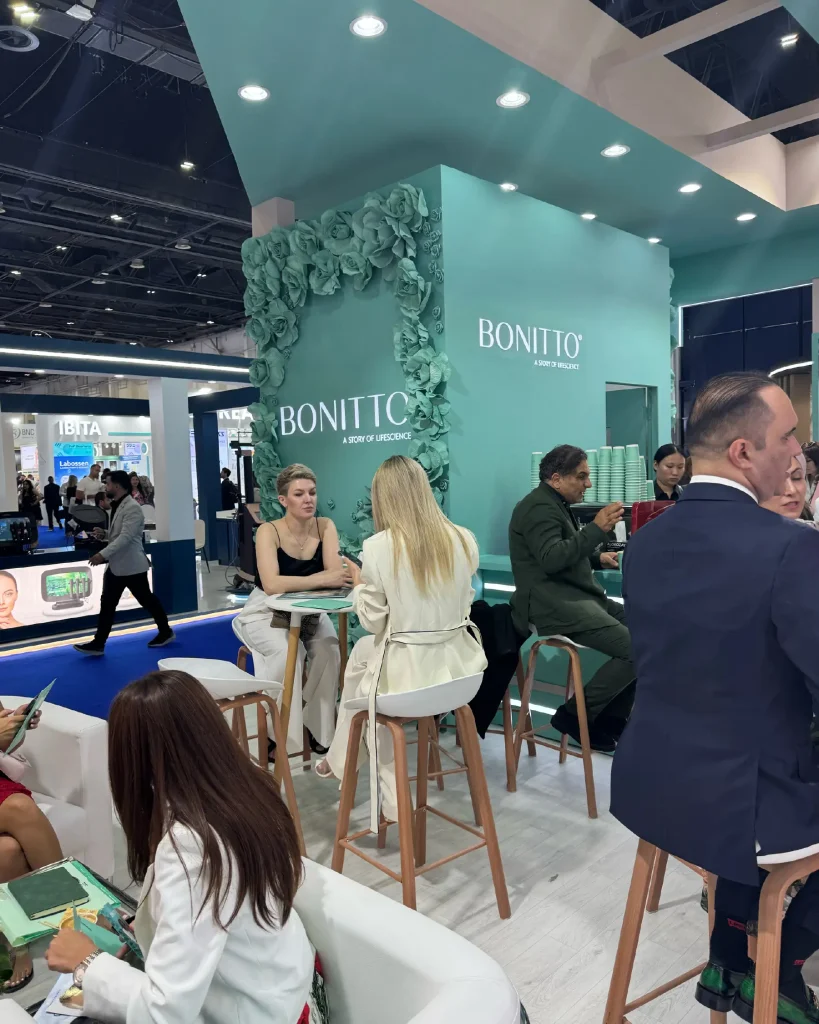 Menschen sitzen und unterhalten sich an einem stilvollen Messestand, der mit grünen Blumenmustern und dem Markennamen „BONITTO“ dekoriert ist. Im Hintergrund sind weitere Stände und Besucher der Messe zu sehen.