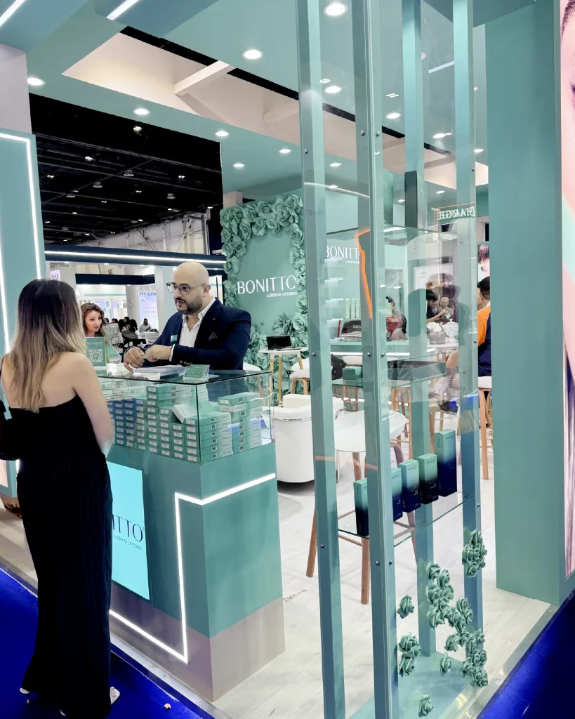 Ein Mann und eine Frau unterhalten sich an einem modernen, türkisfarbenen Messestand mit Glasfronten und Blumenschmuck. Im Stand sind mehrere Personen zu sehen, auf dem Tresen sind Kosmetikprodukte ausgestellt.