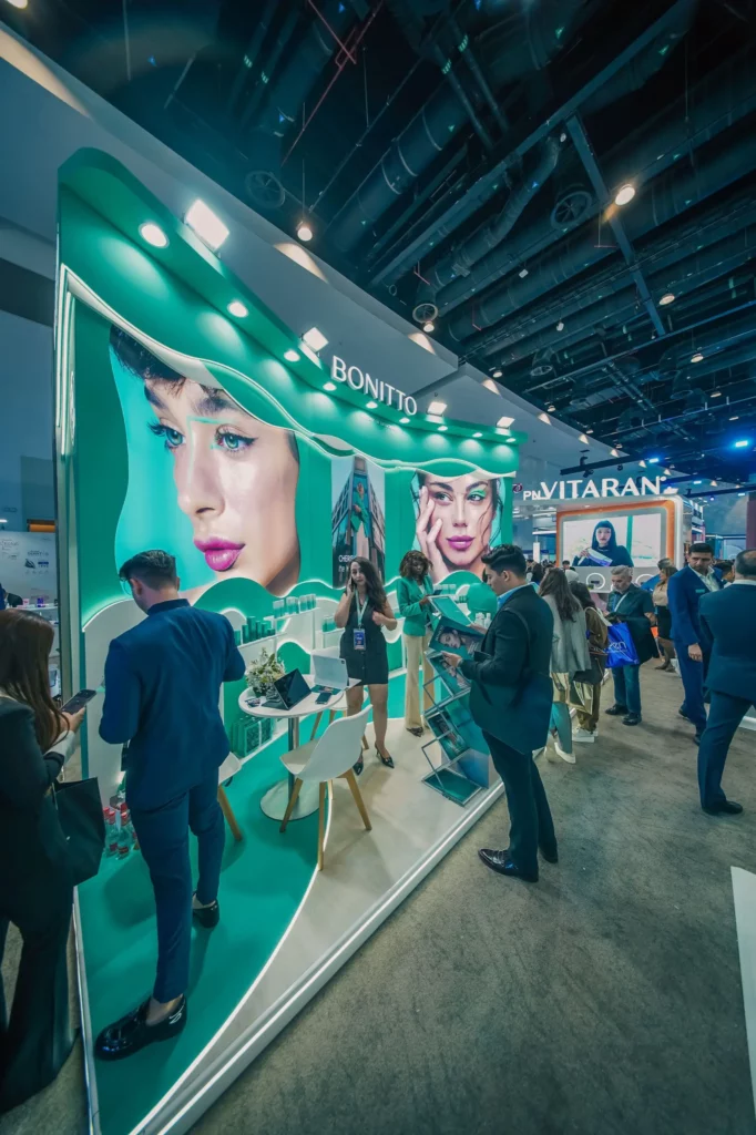 Des personnes interagissent sur un stand d'exposition moderne, avec des images vivantes de visages de femmes sur les panneaux d'affichage. Le stand porte la marque "BONITTO" et présente des accents turquoise, des chaises et une table, dans un centre de convention très fréquenté.