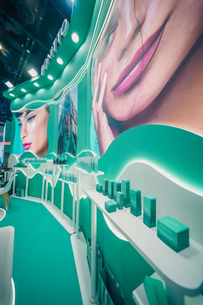 Une cabine de présentation de produits cosmétiques sarcelle et blanche avec des étagères de produits et de grandes images d'un visage de femme maquillée sur le mur arrière incurvé. Le mot "BONITTO" apparaît au-dessus du présentoir.