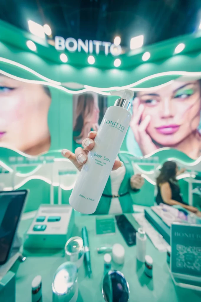 Une personne brandit un flacon blanc de produit de soin de la peau Bonitto dans un stand turquoise vibrant, avec des produits de beauté et du matériel promotionnel exposés sur le comptoir et de grandes images de mannequins en arrière-plan.