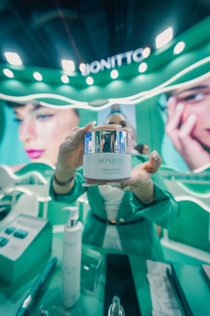 Une personne en tenue sarcelle tient un pot de cosmétique BONITTO Fresh Spirit en direction de l'appareil photo dans un stand de beauté au thème vert et vibrant, avec de grandes images de visages en arrière-plan.