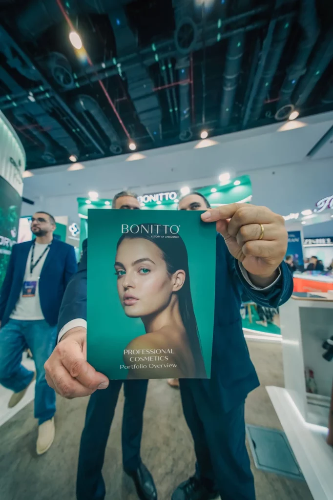 Une personne en costume brandit une brochure de produits cosmétiques BONITTO représentant le visage d'une femme lors d'un événement intérieur animé, avec des personnes et des stands en arrière-plan.
