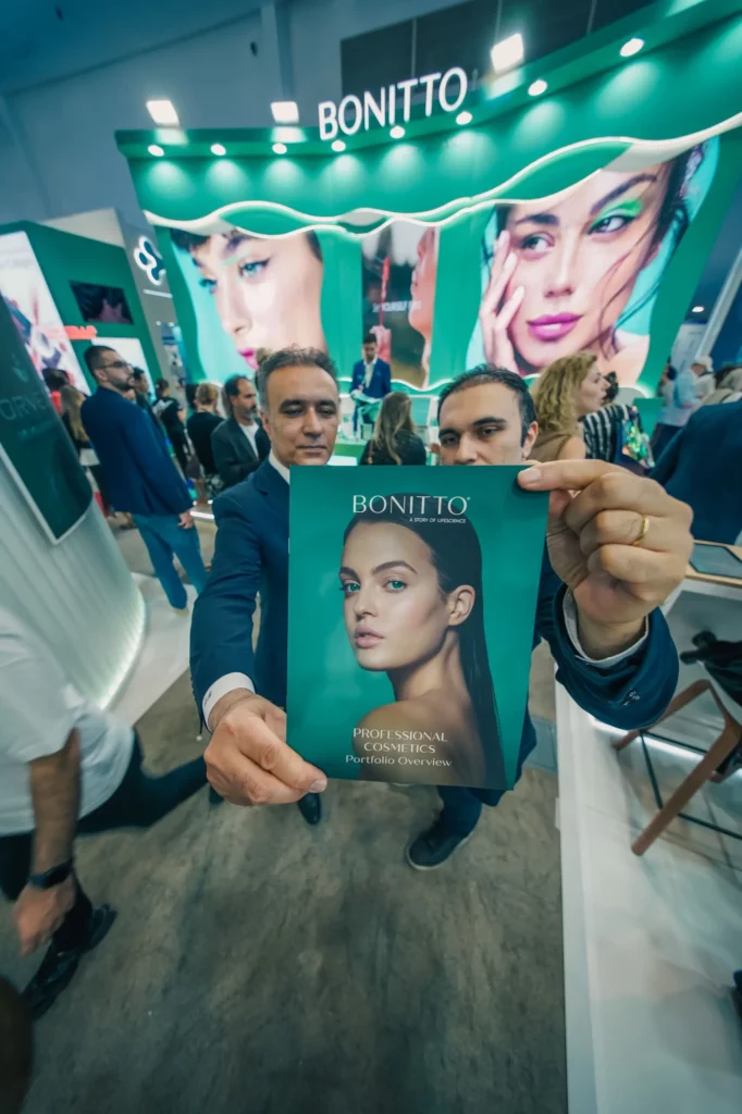 Dans un stand d'exposition très fréquenté, deux hommes brandissent un magazine BONITTO présentant un mannequin au maquillage saisissant. Le stand derrière eux affiche de grandes images du même mannequin et le nom de la marque en lettres blanches lumineuses.