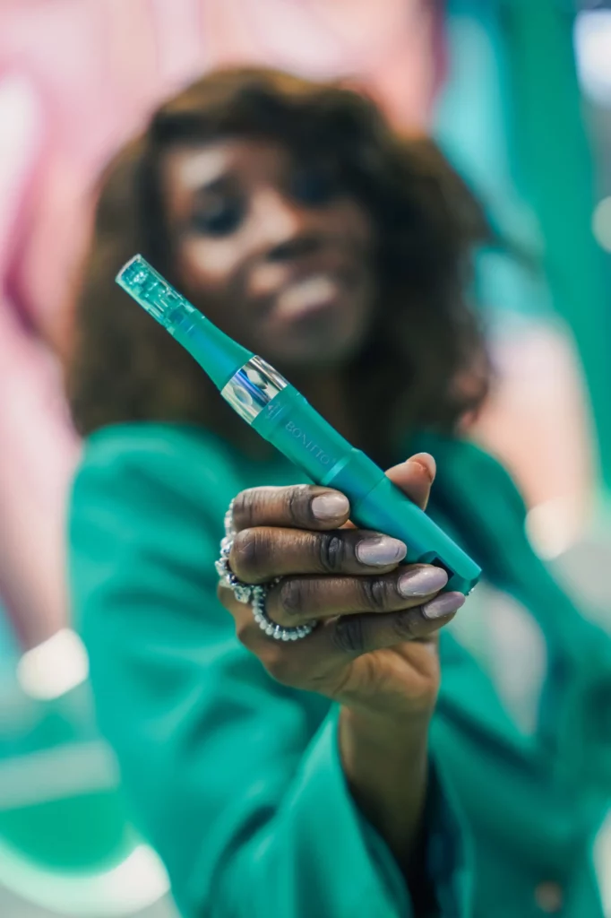 Une femme vêtue d'une tenue sarcelle tient un appareil en forme de stylo sarcelle en direction de l'appareil photo, son visage étant flou à l'arrière-plan. L'appareil est le point central de l'image.
