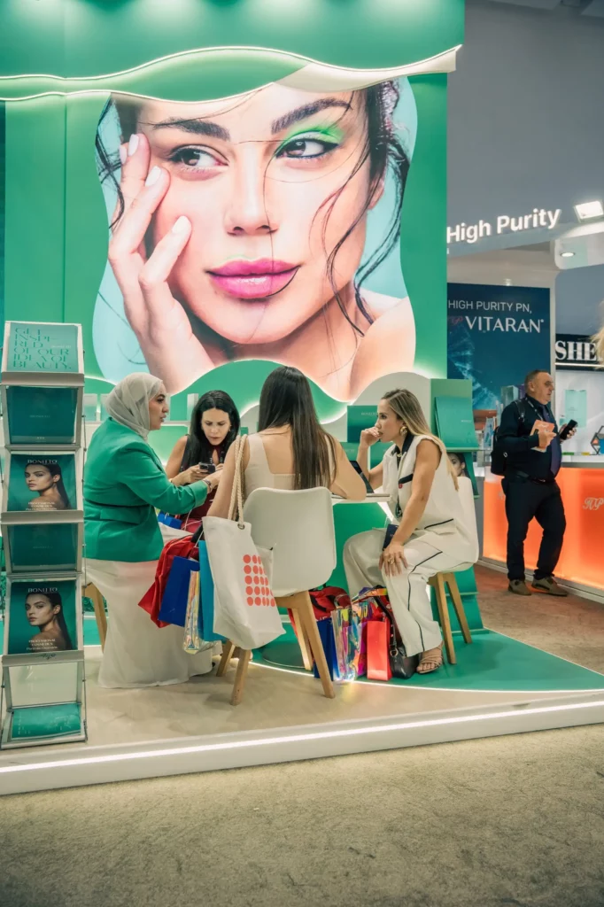 Quatre femmes sont assises autour d'une petite table, discutant et tenant des sacs de courses dans un stand très éclairé avec un grand poster représentant un visage de femme en arrière-plan. Un homme se tient à l'arrière-plan, près d'étagères.