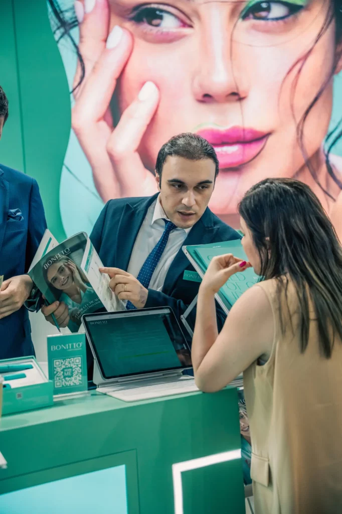 Un homme en costume s'entretient avec une femme à un stand, tenant des brochures. Un grand poster représentant le visage d'une femme se trouve à l'arrière-plan. Un ordinateur portable et du matériel promotionnel sont posés sur le comptoir.