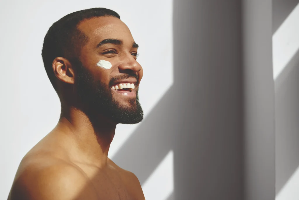 A smiling man with a beard has a dab of cream on his cheek. He is standing against a white background with diagonal shadows.