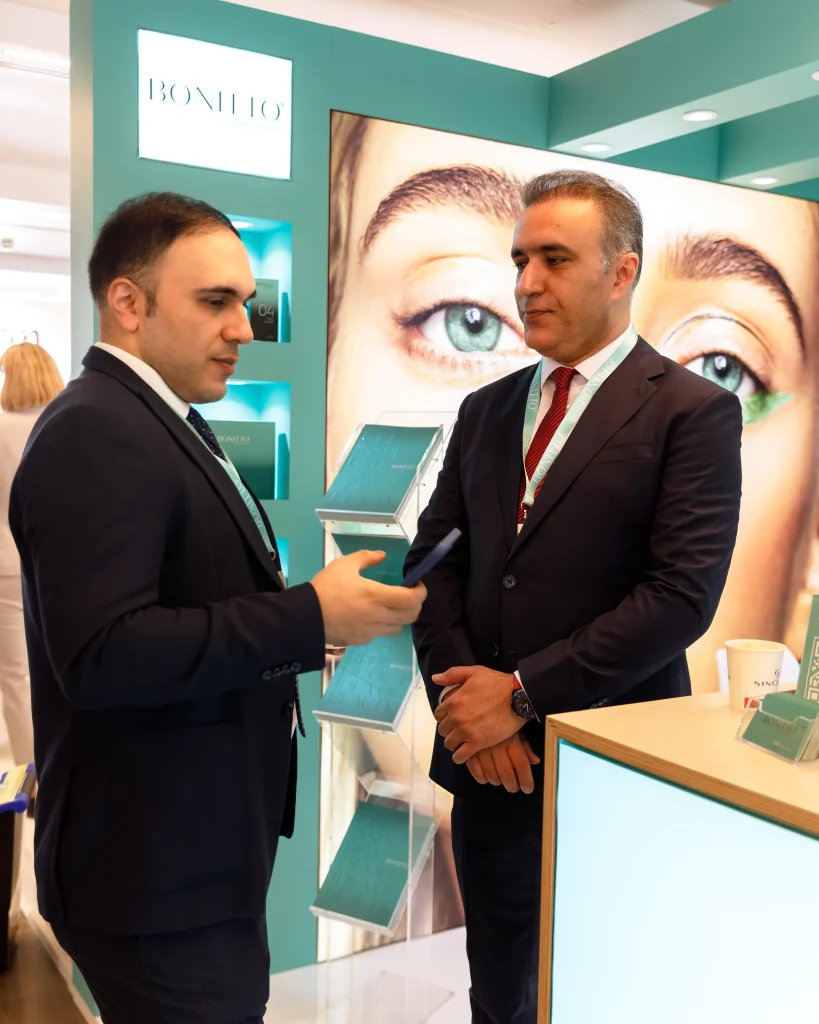 Takım elbiseli iki adam, arka planda büyük bir göz ve kaş resmi bulunan bir ticaret fuarı standında sohbet ediyor. Standda "BONITO" ismi yer alıyor ve turkuaz iç mekanlar ve ürün standları bulunuyor.