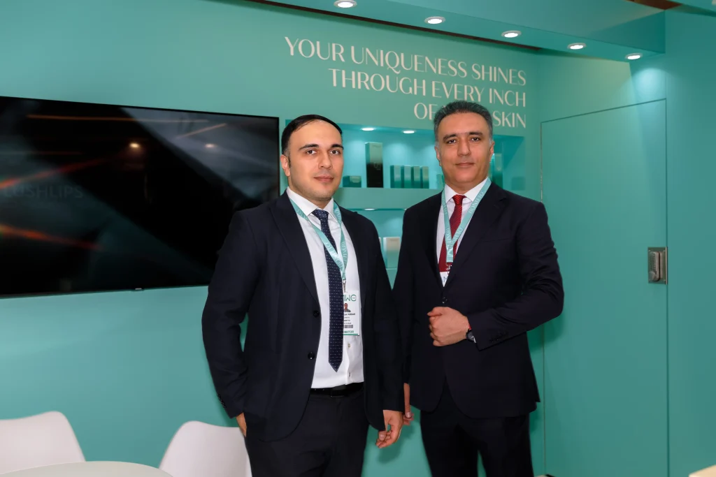 İş takım elbiseli iki adam, üzerinde "Benzersizliğiniz cildinizin her santiminden parlıyor" yazan, turkuaz renkli bir arka plana sahip bir stantta fotoğraf çektirmek için duruyor. Arkalarında bir televizyon ve ürünlerin bulunduğu raflar var.