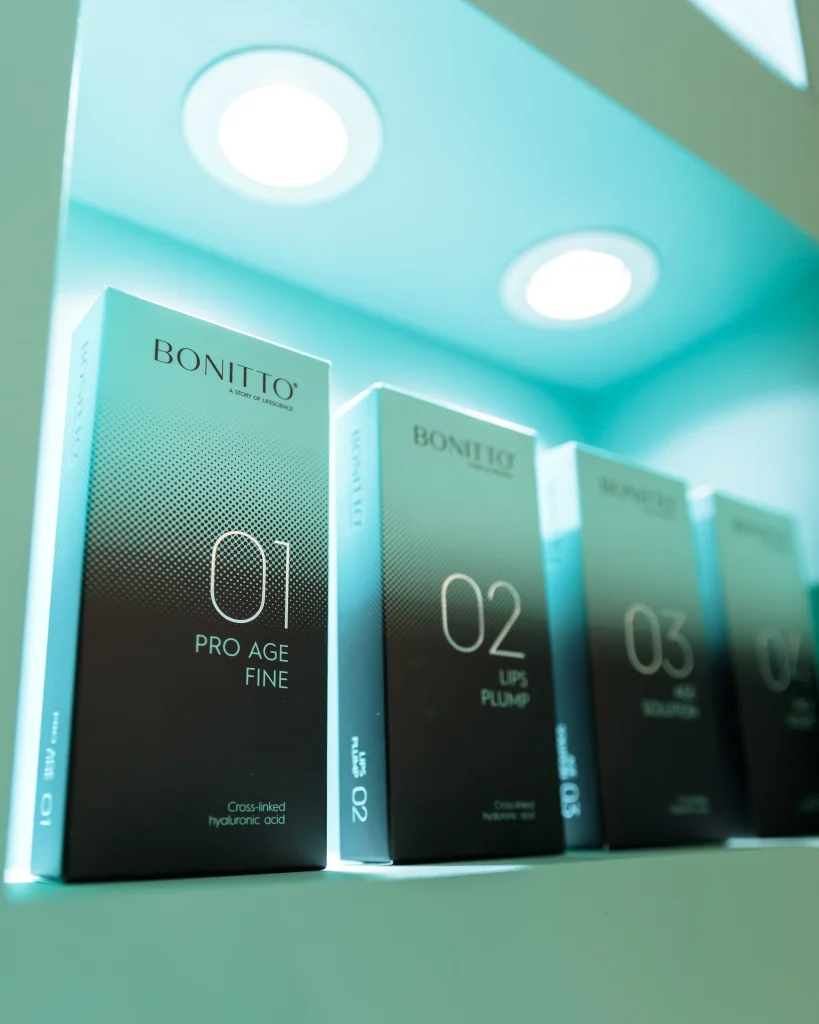 Üzerinde 01 Pro Age Fine, 02 Lips Plump ve 03 Hair Volumizer yazan Bonitto cilt bakım ürünleri kutuları, turkuaz renkli aydınlatmanın altında bir rafta dik olarak sergileniyor.