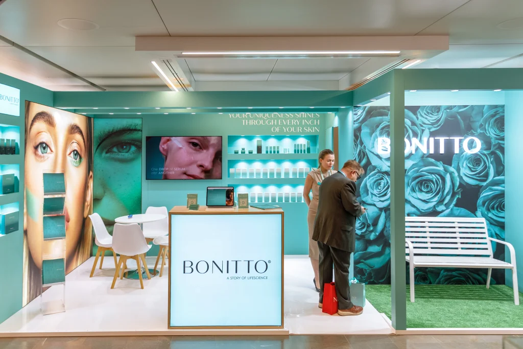 BONITTO" etiketli modern bir cilt bakımı standında ürün teşhirleri, büyük yüz ve turkuaz gül resimleri, beyaz bir bank, sandalyeler ve parlak, şık ışıklandırma altında tezgahın yakınında etkileşimde bulunan iki kişi yer alıyor.