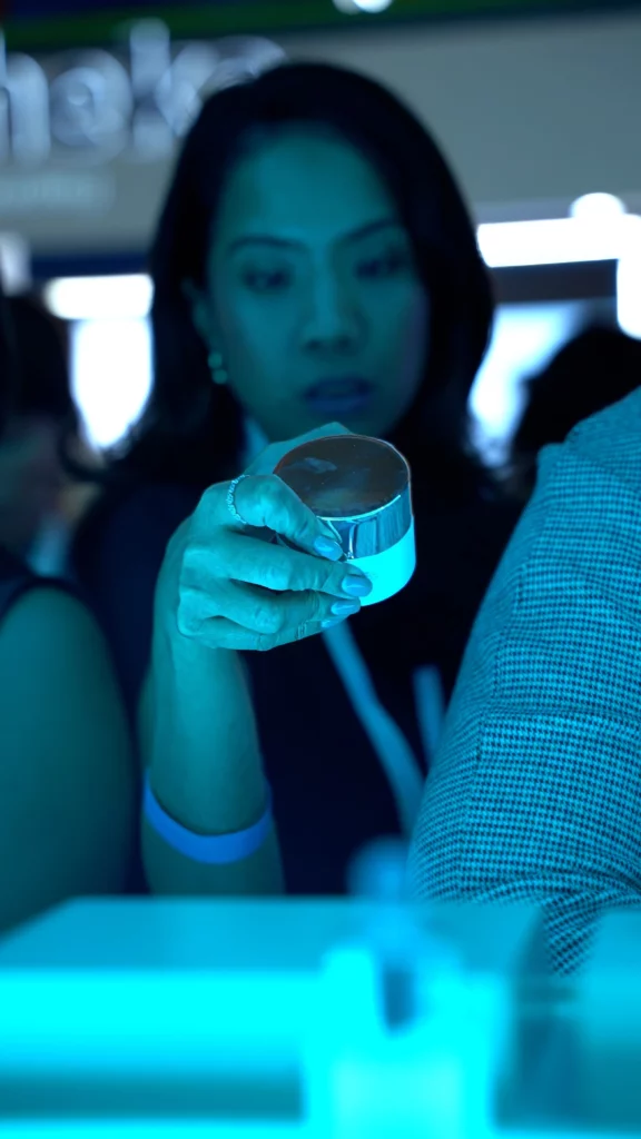 A woman holds a small round container, possibly of cream or cosmetics, focusing intently on it. The image is tinted blue, with other people blurred in the background.