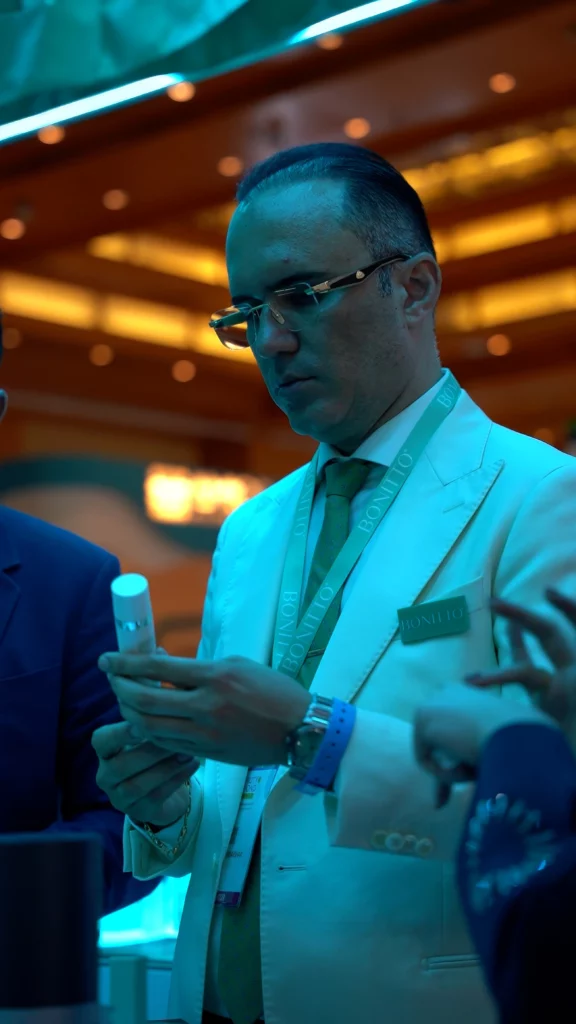 A man in a white suit and glasses examines a small cylindrical object under blue lighting at an indoor event, with blurred people and warm lights in the background.