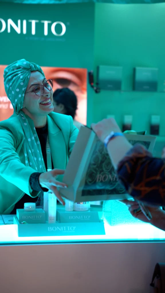 A smiling woman in a green blazer and polka-dot headscarf hands a boxed product to a customer at a brightly lit, modern retail counter. Skincare products and branding labeled "BONITO" are visible on the counter.