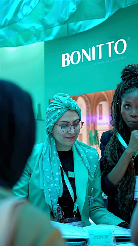 Two women stand at a booth with a "BONITTO" sign behind them. One wears glasses, a polka-dot headscarf, and a blazer, while the other has long braided hair and holds a pen. The lighting is blue-green.