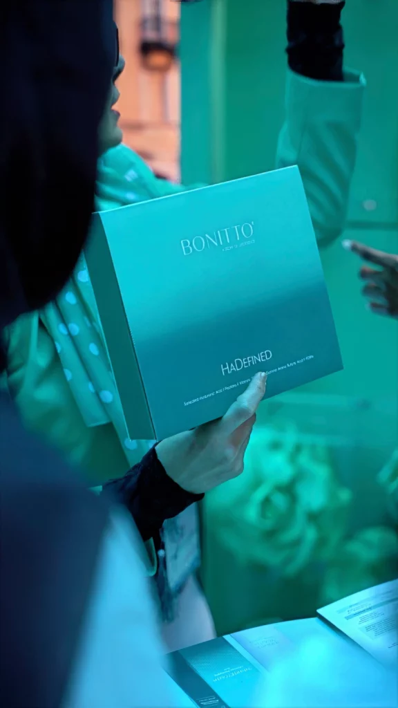 A person holds a teal BONITTO box labeled "HADFINED" in a brightly lit, blue-green environment, surrounded by other people.
