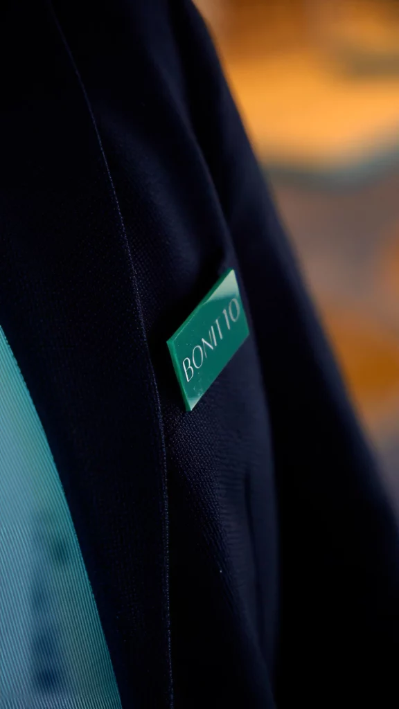 Close-up of a dark-colored blazer with a green name tag on the lapel. The tag has the word "BONITO" written in white letters. The background is softly blurred.