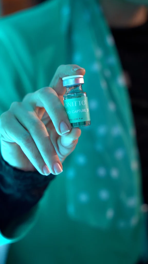 A person holding a small vial labeled "NITIQ CO CAPTURE 5" between their thumb and forefinger, with a teal-colored background and the person's clothes visible in soft focus.