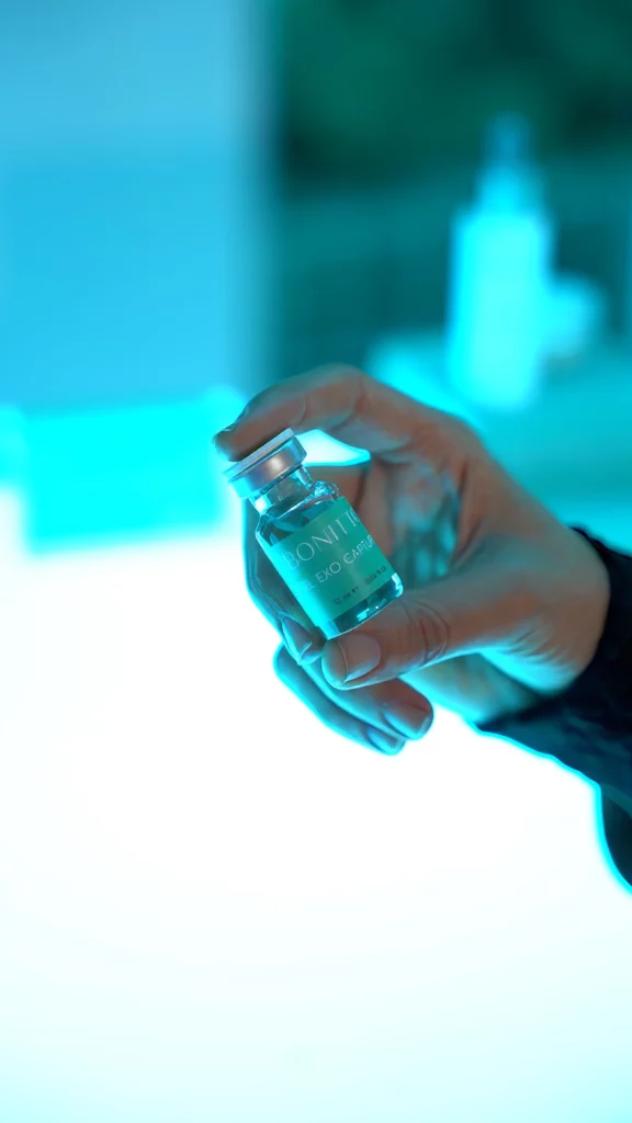 A hand holding a small clear vial labeled "COVID-19 VACCINE" against a bright blue and white background.