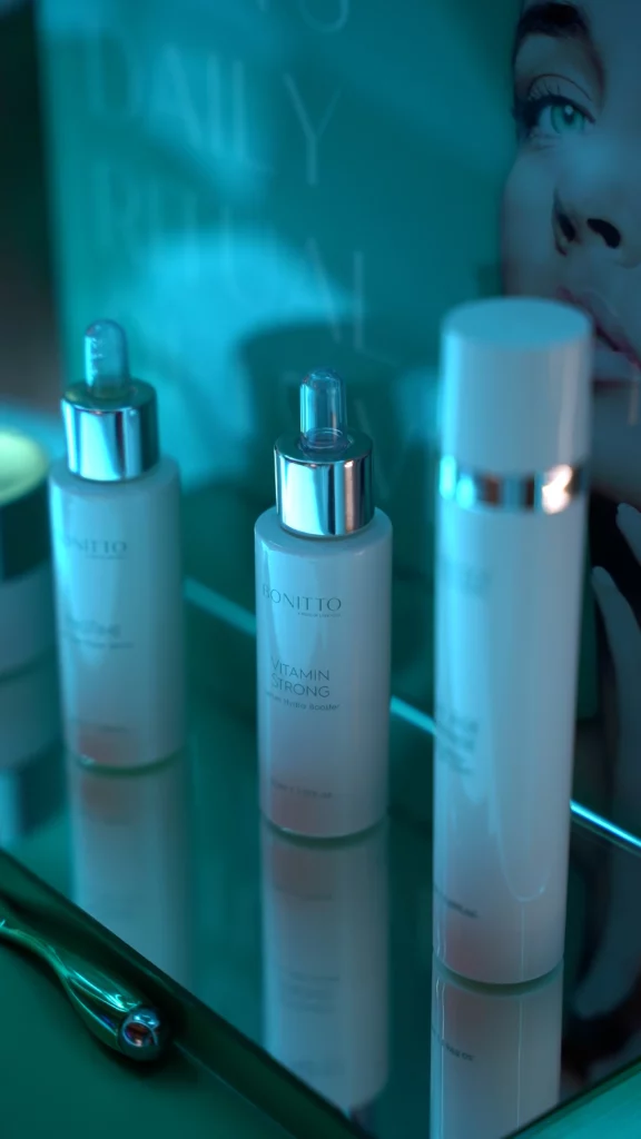 Three skincare product bottles, including a dropper bottle labeled "Vitamin Strong," are displayed on a reflective glass surface with a blurred poster of a woman's face in the background under teal lighting.