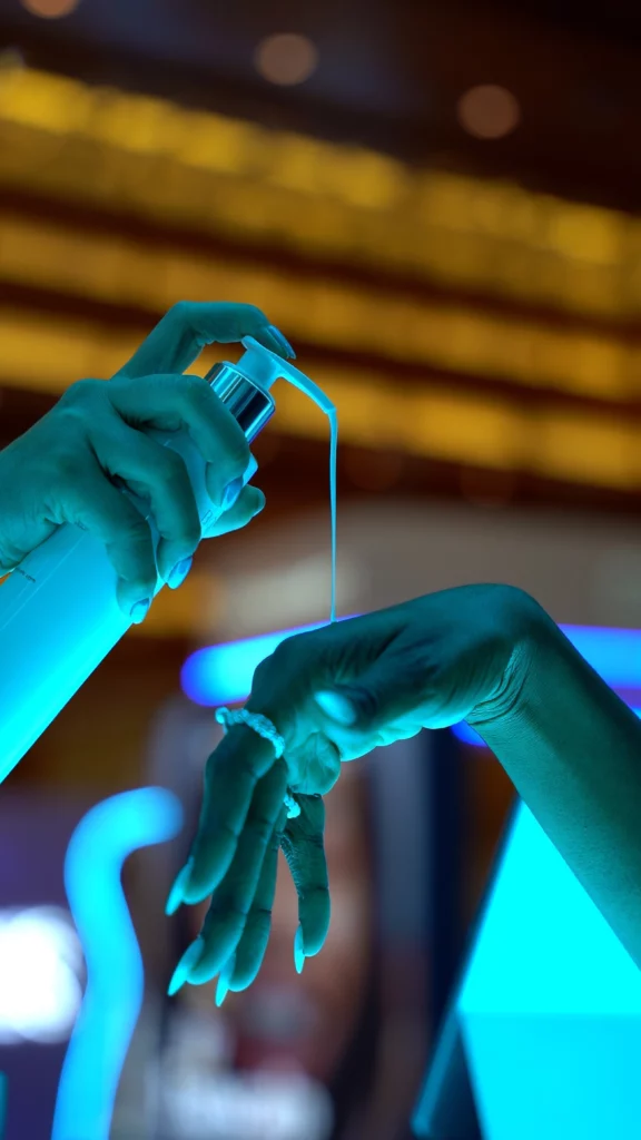 A close-up of one hand dispensing lotion from a pump bottle onto the back of another hand, both illuminated by a blue light in a modern, blurred background.