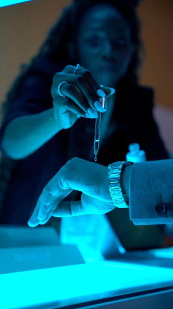A person wearing a watch extends their hand while another person applies liquid from a dropper onto their wrist under blue lighting.