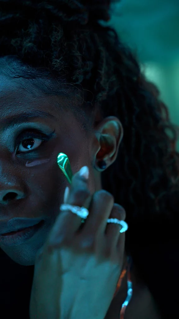 A person with curly hair applies a green under-eye patch to their face. Their hand, adorned with rings, gently touches their cheek. The image has a cool, blue-toned lighting.