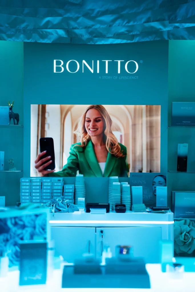 A display for BONITTO skincare features a smiling woman in a green blazer taking a selfie on a large screen, surrounded by neatly arranged skincare products and a blue-toned modern setup.