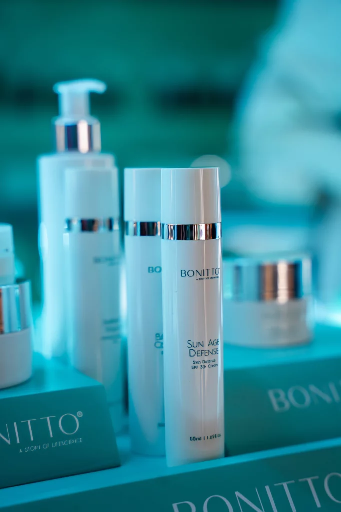 Several white skincare product bottles and jars with silver accents are displayed on a turquoise surface. The visible label reads "BONITTO SUN AGE DEFENSE." The background is blurred, highlighting the products.
