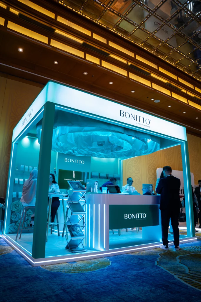 A modern, illuminated exhibition booth labeled "BONITTO" with glowing blue lights and people interacting inside and around the booth, set in a large, upscale indoor venue with a high ceiling.