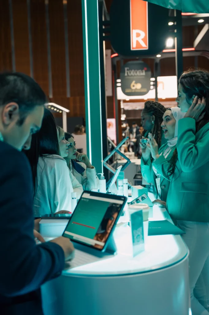 People interacting at a modern exhibition booth with digital tablets and product displays. Several women stand and talk, while a man writes on a tablet. Glowing blue lights illuminate the scene.