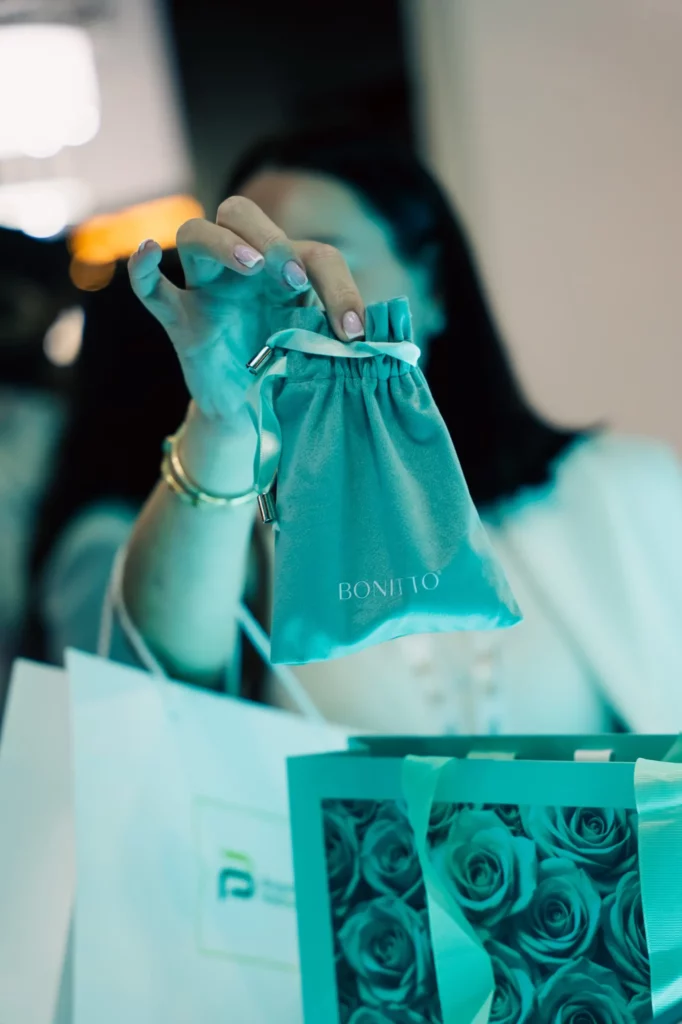 A person holds up a small drawstring pouch labeled "BONITO" in front of shopping bags, one of which features an image of roses. The person's face is out of focus, and only their hand and the pouch are clear.