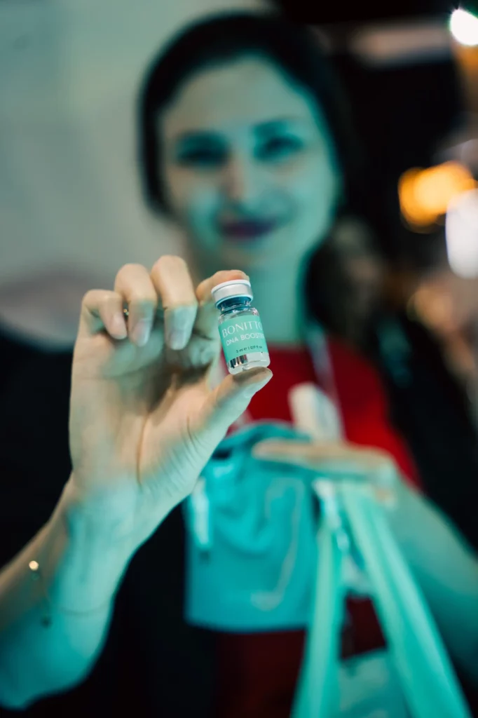 A woman holds a small vial labeled "BONITTO" toward the camera, with the vial in sharp focus and her face blurred in the background. She has painted nails and wears a red top and a lanyard.