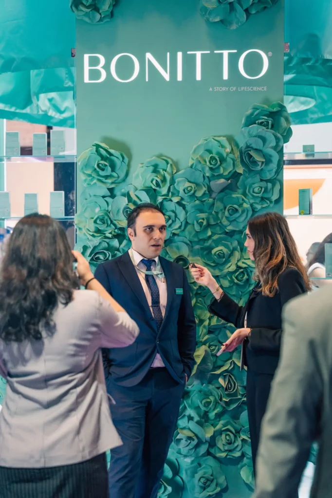 A man in a suit stands in front of a green floral display with the word “BONITTO” above him, while two women interact with him; one is interviewing and another is taking a photo.