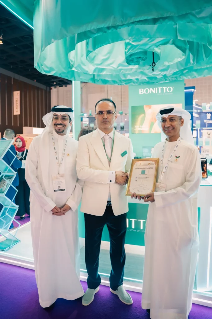 Three men in white attire stand together at an exhibition booth. One man in the center shakes hands with the man on the right, who holds a framed certificate. Colorful displays and the "BONITTO" sign appear in the background.