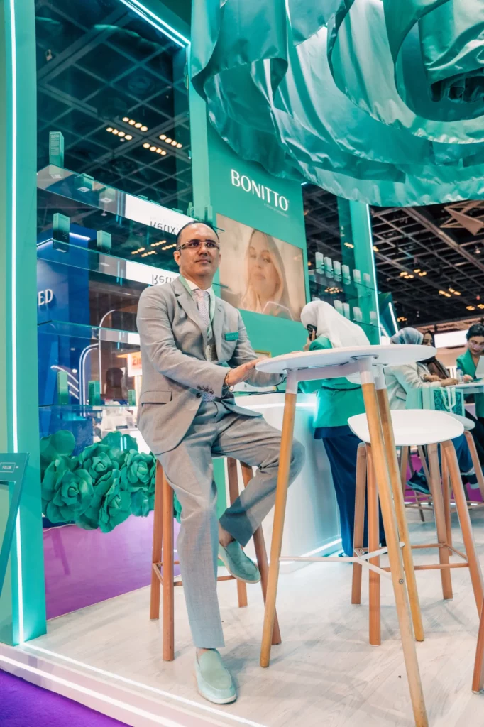A man in a light gray suit sits on a high chair at a modern exhibition booth with a "BONITTO" sign, turquoise decor, and green floral accents. People work and interact in the background.