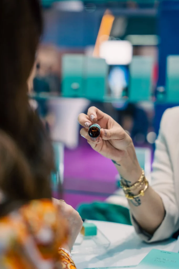A person wearing a white blazer and gold bracelets holds up a gemstone for another person to examine in a brightly lit setting with a blurred, colorful background.
