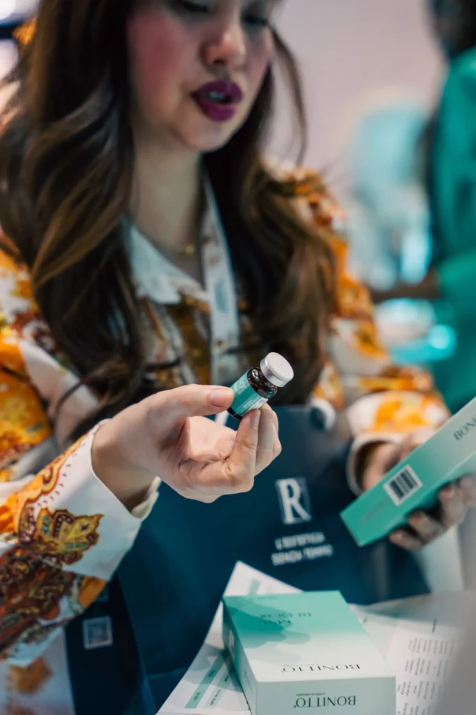 A woman in a patterned shirt holds a small vial and a teal box, possibly discussing or presenting the product. She appears engaged and focused, with several product boxes visible on the table in front of her.
