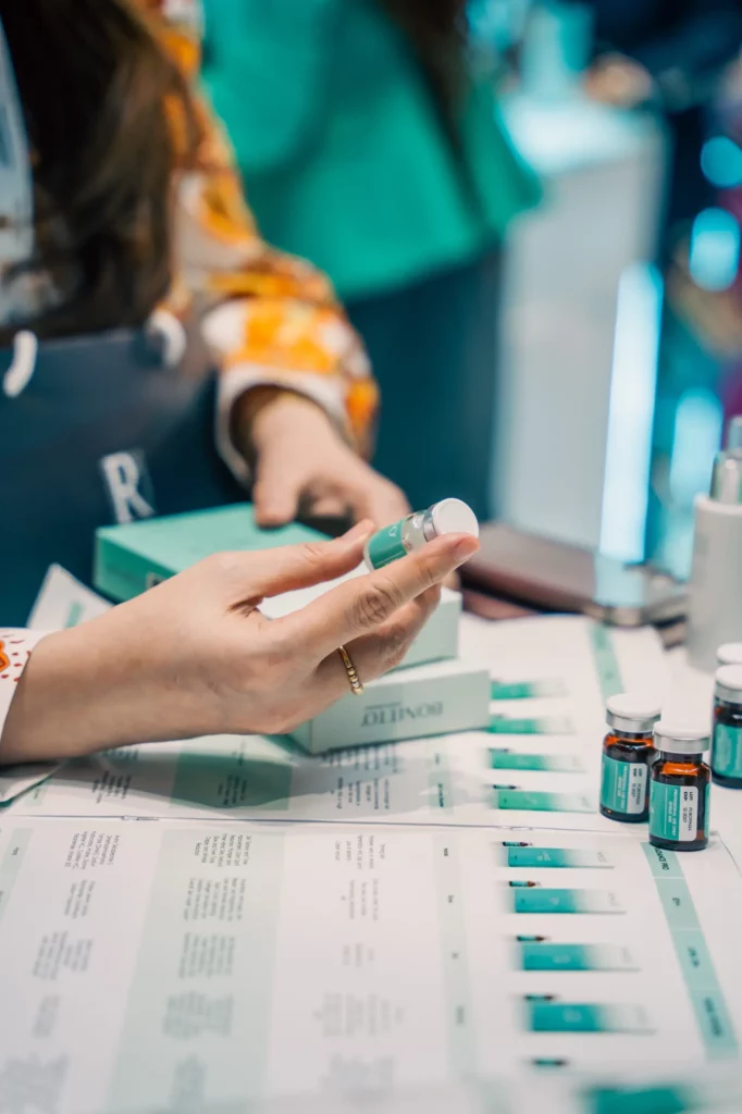 A person holds a small vial while examining its label. Several more vials and boxes are arranged on a table covered with informational sheets. The scene suggests a medical or cosmetic setting.