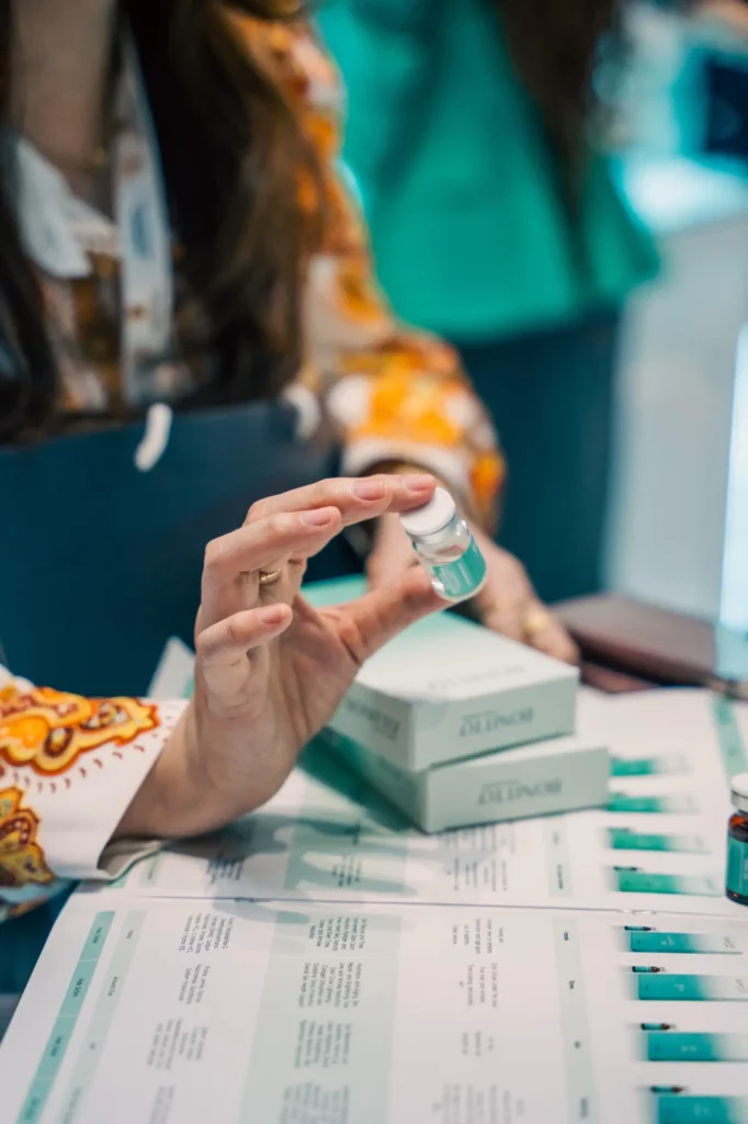 A person with long hair wearing a patterned sleeve holds a small clear vial above boxes and pamphlets on a table, possibly at an event or display related to skincare or medical products.