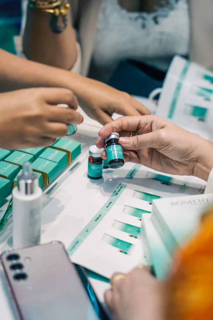 People’s hands holding and examining small cosmetic serum bottles, with more beauty products, sample packaging, and a smartphone on a table. The scene suggests a skincare or cosmetics consultation.