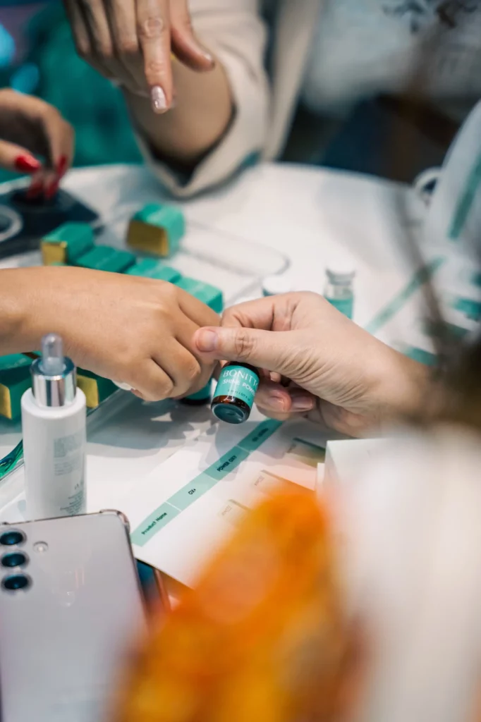 A person applies a cosmetic product to another person’s hand at a table with skincare products, bottles, and a smartphone nearby.