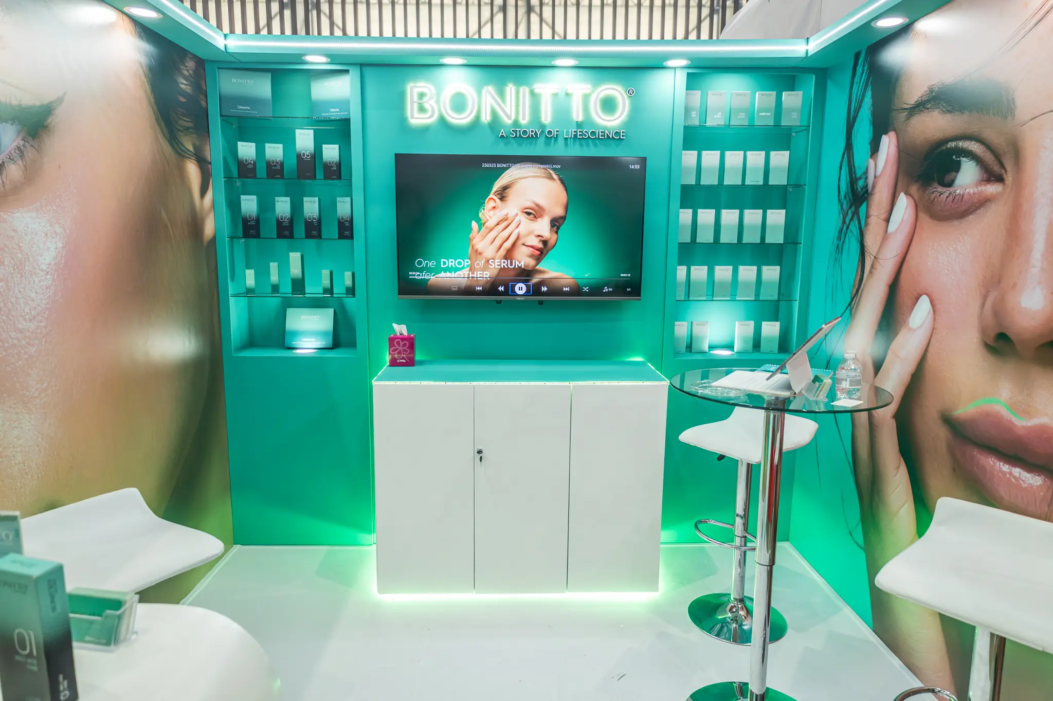 كشك لمستحضرات التجميل بجدران خضراء يعرض مستحضرات BONITTO للعناية بالبشرة. أرفف تعرض المنتجات وشاشة كبيرة تعرض وجه امرأة و"قطرة واحدة من السيروم". توجد صورتان عن قرب لوجه امرأة على جانبي الكشك.