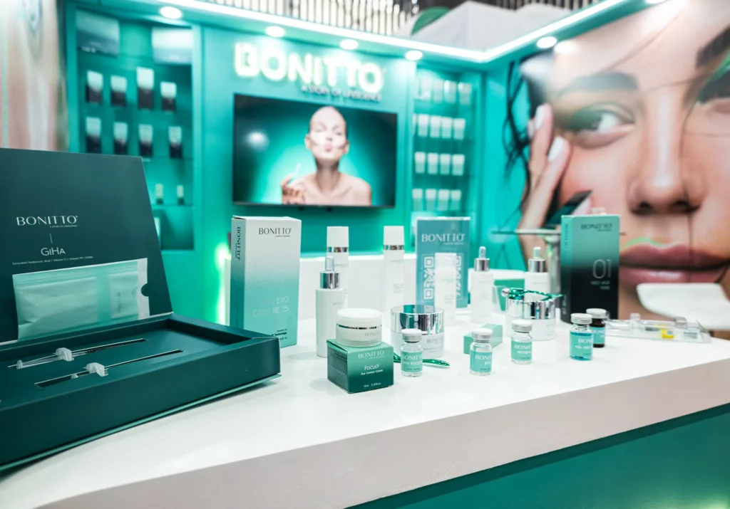 عرض لمنتجات بونيتو للعناية بالبشرة مرتبة على منضدة بيضاء، مع برطمانات وزجاجات وصناديق. وتظهر خلفية باللون الأزرق المخضر على أرفف المنتجات وصورة لوجه امرأة ذات بشرة متوهجة وصحية.