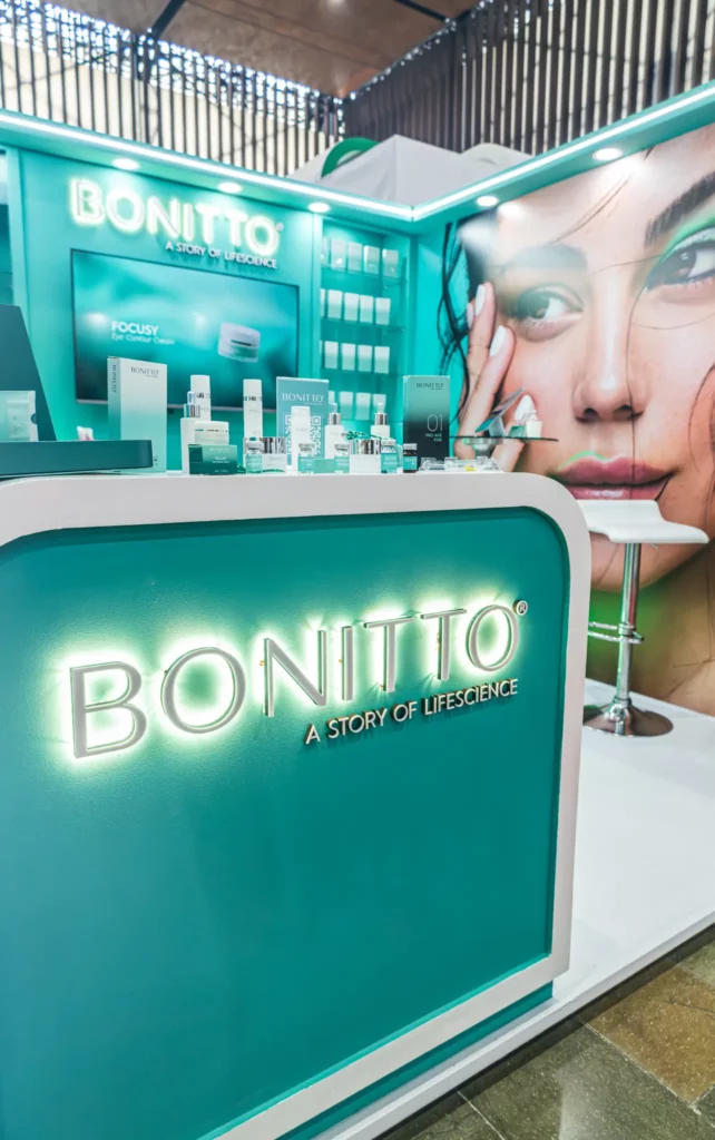 كشك BONITTO للعناية بالبشرة بلون البط البري مع منتجات معروضة على المنضدة ولافتات تحمل العلامة التجارية وملصق كبير لوجه امرأة في الخلفية. الشعار مكتوب عليه "قصة حياة.