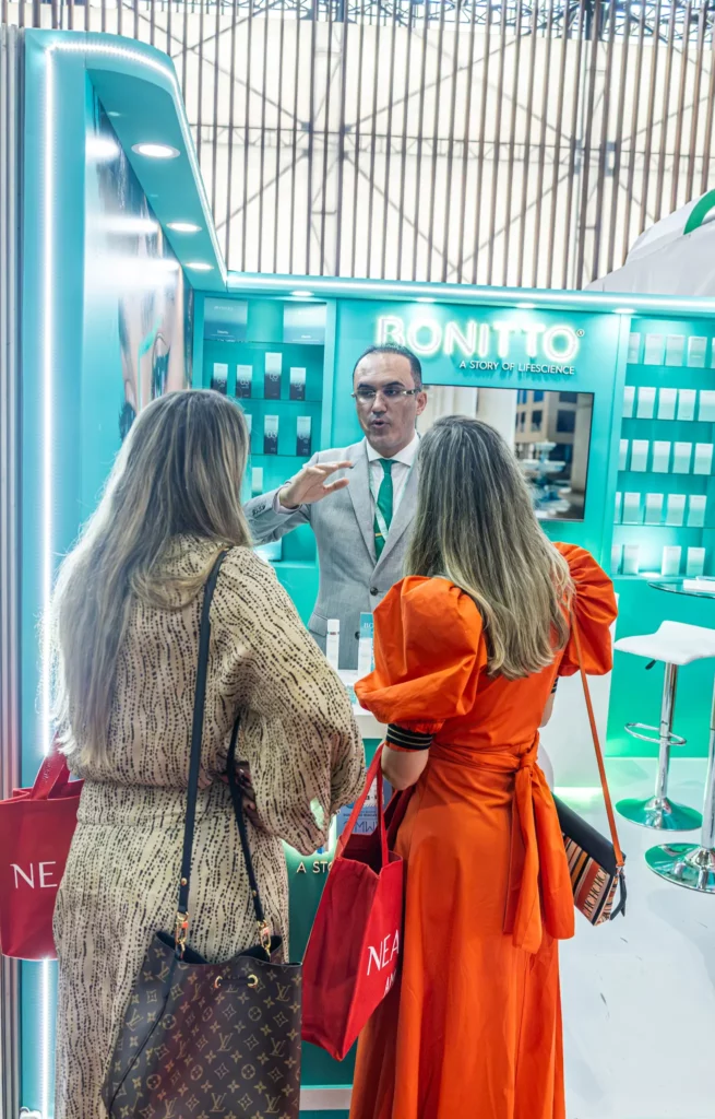 رجل يرتدي بدلة رسمية يتحدث إلى امرأتين تحملان حقيبة يد حمراء في كشك عرض BONITTO المضاء بإضاءة ساطعة، مع جدران مخضرة ورفوف من المنتجات في الخلفية.