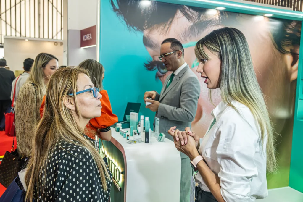 امرأتان تتحدثان بالقرب من شاشة عرض لمنتجات العناية بالبشرة في معرض تجاري، بينما يتفاعل رجل يرتدي بدلة مع آخرين في الكشك. وتظهر في الخلفية صورة كبيرة لوجه والعديد من مستحضرات التجميل.