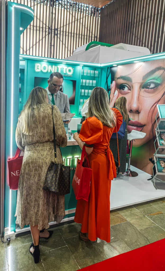تقف ثلاث نساء يحملن أكياساً حمراء في كشك BONITTO للعناية بالبشرة مضاء بشكل ساطع، ويتحدثن إلى رجل خلف المنضدة. يتميز الكشك بصورة كبيرة لوجه امرأة ورفوف تعرض المنتجات.
