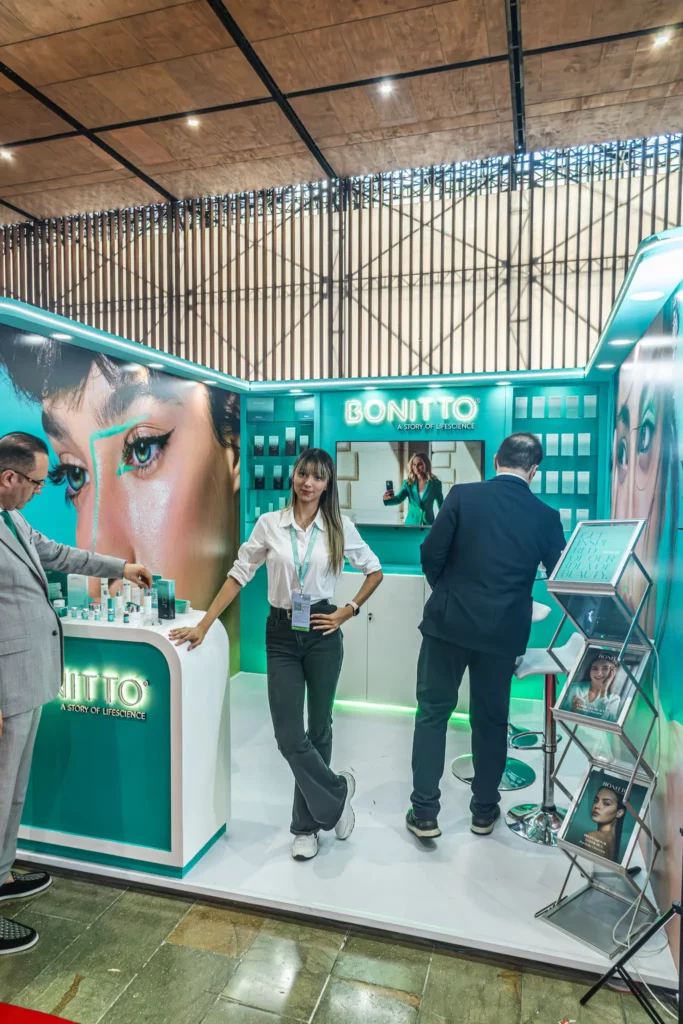 امرأة تقف أمام كشك BONITTO للعناية بالبشرة مضاء بشكل ساطع في أحد المعارض، مع وجود رجلين يتفاعلان بالقرب منها ومنتجات ترويجية وصور كبيرة لوجوه معروضة حول الكشك.